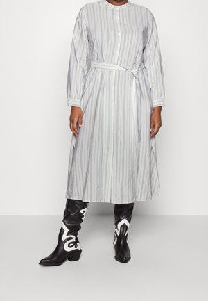 Pruhované šaty s dlouhým rukávem ve světle šedé barvě s tmavšími vertikálními pruhy, stáhnuté v pase páskem, doplněné černými kovbojskými botami s bílými akcenty.