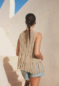 Béžový háčkovaný top bez rukávů s diamantovými vzory a třásněmi na spodním lemu, spárovaný se světle modrými denimovými šortkami na pozadí bílé zdi.