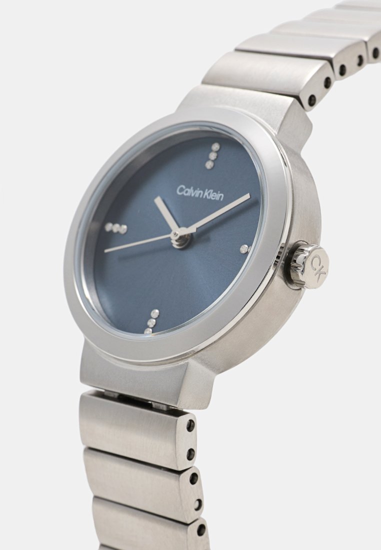 RVS-horloge met een ronde blauwe wijzerplaat, zilverkleurige uurmarkeringen en een metalen schakelarmband. Voorzien van een kroon met "CK" gravure.