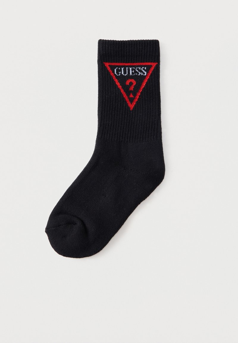 Chaussette noire côtelée avec écusson triangulaire rouge du logo Guess affichant un texte blanc « GUESS » et un point d’interrogation rouge au niveau du mollet.