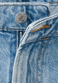 Tela de mezclilla azul claro con textura visible, con cierre de cremallera y un botón metálico plateado con relieve que dice "MNG DENIM". Detalles en costuras marrones.