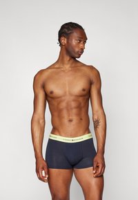 Sorte bomullsboksershorts med et lysegrønt elastisk midjebånd som har "Tommy Hilfiger"-logoen. Modellen står sideveis for å vise passformen.