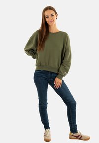 Sudadera oversized de color verde oliva con puños y dobladillo acanalados, combinada con vaqueros ajustados de color azul oscuro y zapatillas beige con rayas burdeos.