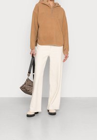 Pullover beige in pile con collo a zip, abbinato a pantaloni bianchi a gamba larga e una borsa a quadri marrone. Tessuto testurizzato, vestibilità rilassata e dettagli a cerchio.