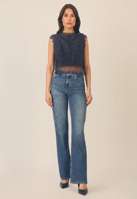 Top corto in pizzo blu navy con orlo scallop e motivo floreale, abbinato a jeans a campana blu chiaro e tacchi a punta neri.