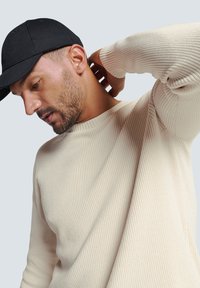 Beige geribbelde trui met een ronde hals; zwarte pet; textuur benadrukt door het breipatroon; zachte, relaxte pasvorm.