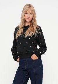 Sweatshirt noir avec des motifs de cœurs dorés, col rond et poignets côtelés. Assorti à un pantalon en denim bleu foncé. Texture de tissu douce.