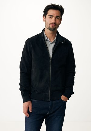 Chaqueta bomber de ante negra con cierre de cremallera en el frente, puños y cuello acanalados. Combinada con jeans azul oscuro, sobre un fondo neutro.