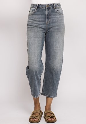 Frau trägt hoch taillierte, ausgewaschene blaue, kurz geschnittene Jeans und hellbraune Sandalen mit Nieten, steht vor einem schlichten hellen Hintergrund.