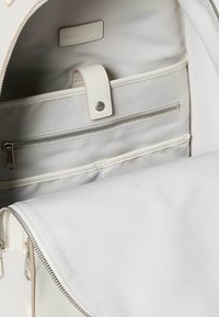 Interiér bílého látkového batohu se šedou podšívkou, vybavený kapsou na zip a zapínáním na druk. Na štítku je nápis "Calvin Klein Jeans."