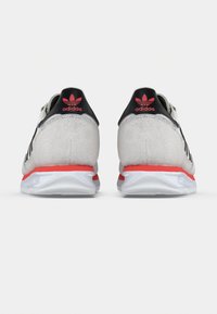 Szürke velúr tornacipők fekete részletekkel, piros sarokrészlettel és fehér talppal. Az Adidas logó látható a sarokrészen.