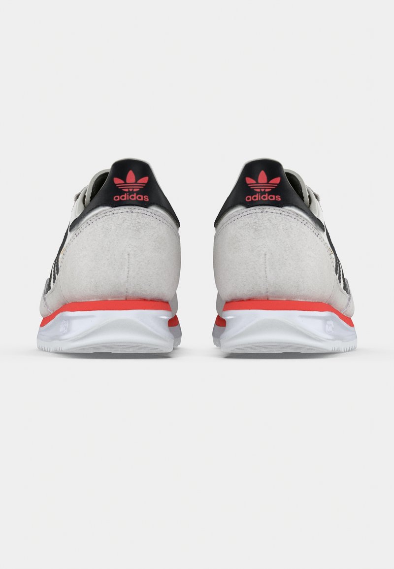 Baskets en daim gris avec des accents noirs, dotées d'un détail rouge au talon et d'une semelle blanche. Le logo Adidas est visible sur le talon.