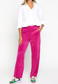 Pantaloni a gamba larga in velluto a coste rosa con una texture morbida, abbinati a una camicia bianca con bottoni e sneakers colorate. Presentano tasche laterali.