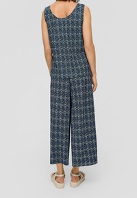 Combinaison sans manches bleu marine avec un motif géométrique, une coupe à jambes larges et une texture de tissu douce, portée avec des sandales beiges.