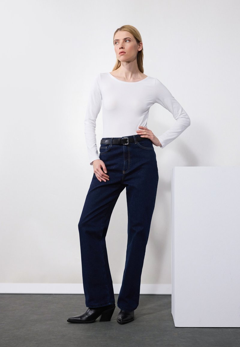 Blusa blanca de manga larga combinada con vaqueros de tiro alto de color azul oscuro. Los vaqueros tienen un corte recto y un cinturón negro con una hebilla plateada.