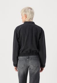Svart denim bomberjacka med elastisk midja, ribbad manschetter och krage. Bärs med grå denimjeans som har bakfickor.