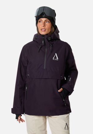 Femme portant une veste de ski violet foncé avec capuche et un pantalon beige, avec des lunettes de ski noires sur la tête, regardant sur le côté.