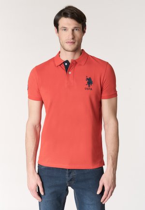 Uomo con polo rossa USPA con logo blu navy, in piedi contro uno sfondo chiaro uniforme, mani rilassate lungo i fianchi.
