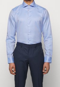 Camicia azzurra chiara con un motivo sottile, maniche lunghe e colletto a punte. Abbinata a pantaloni blu scuro. Chiusura frontale con bottoni.