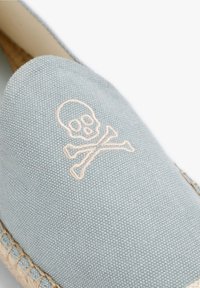 Lichtblauwe canvas instapschoen met een in crèmekleur geborduurde doodskop en kruisbeenderen. Jute afwerking langs de zool voegt textuur toe.