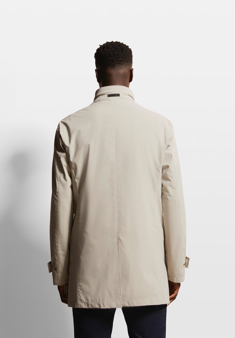 Manteau imperméable beige avec un col montant, des manches longues et une coupe droite. Présente une texture lisse et des détails de couture subtils.