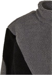 Grå fleecepullover med høj krave, der har en tekstureret, fluffy overflade. Sorte elementer i siderne skaber et kontrasterende design.