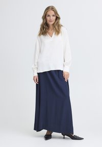 Blusa bianca con scollatura a V e maniche lunghe abbinata a una lunga gonna blu navy; modella che indossa tacchi neri con punta appuntita e dettagli decorativi.