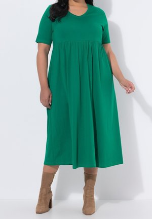 Femme portant une robe midi verte à manches courtes et des bottines en daim beige, debout devant un fond blanc uni.