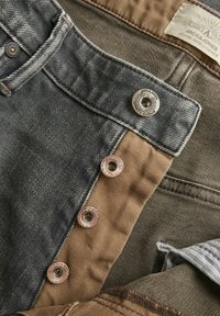 Shorts de mezclilla con un lavado oscuro, botones de cobre, tela beige en contraste y una etiqueta tejida, que muestran una combinación de texturas y colores.