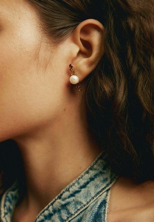 Vista lateral de una persona que lleva un pendiente de oro con una gran perla blanca y pequeñas piedras preciosas rojas, con cabello oscuro y ropa de mezclilla.