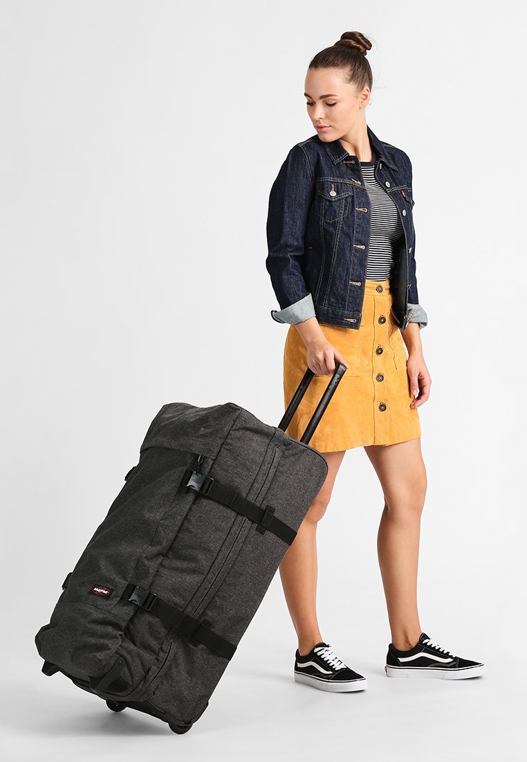Gran bolsa de tela gris de tipo duffel con asas negras y ruedas, siendo tirada por una persona que lleva una chaqueta de mezclilla, una camiseta de rayas y una falda amarilla.