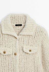 Cardigan tricoté épais écru avec large col, deux poches poitrine à rabat et boutons dorés en forme de nœud.