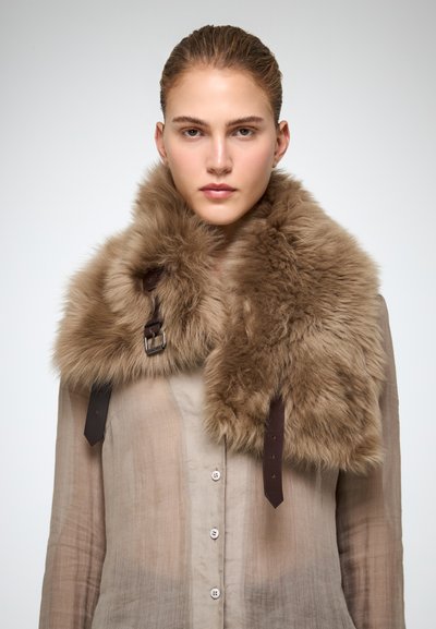 Una donna con i capelli tirati indietro indossa una camicia trasparente beige con bottoni e un grande colletto di pelliccia marrone chiaro con cinturini in pelle scura e una fibbia metallica.