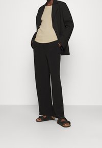 Svart skräddarsydd kavaj över en beige åtsittande topp, kombinerad med svarta vida byxor och svarta sandaler. Slät textur, avslappnad design.