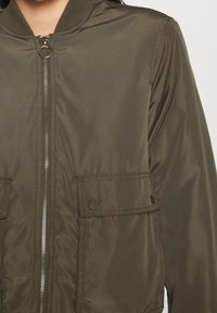 Olive zelená bomber bunda s hladkou, lesklou texturou, předním zipovým uzávěrem a dvěma kapsami na patentky po stranách.