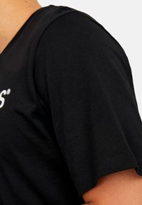 Nahaufnahme einer Person, die ein schwarzes kurzärmliges T-Shirt trägt, auf dem ein Teil des weißen Textes auf der Brust gegen einen einfarbigen Hintergrund sichtbar ist.