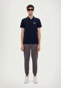 Homem com óculos de sol pretos, camisola polo azul-marinho, calças de treino cinzas e ténis brancos, de pé contra um fundo branco.