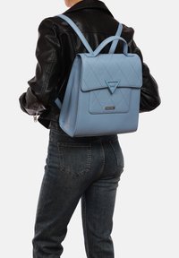 Zaino in pelle blu chiaro con design trapuntato, patta triangolare e spallacci regolabili, indossato da una persona con giacca nera e jeans.