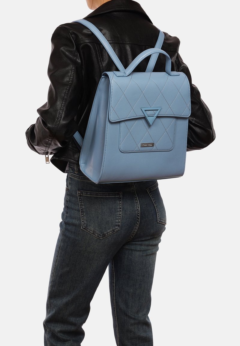 Zaino in pelle blu chiaro con design trapuntato, patta triangolare e spallacci regolabili, indossato da una persona con giacca nera e jeans.