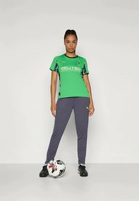 Woman in green sports jersey and gray pants stands with one foot on a black and white soccer ball against a plain background.