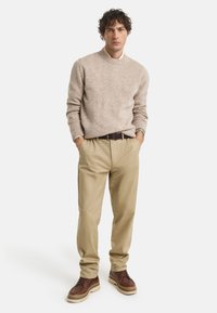 Pull en maille beige, pantalon beige, chaussures marron foncé avec une semelle texturée. Le mannequin se tient avec les mains dans les poches devant un fond blanc.