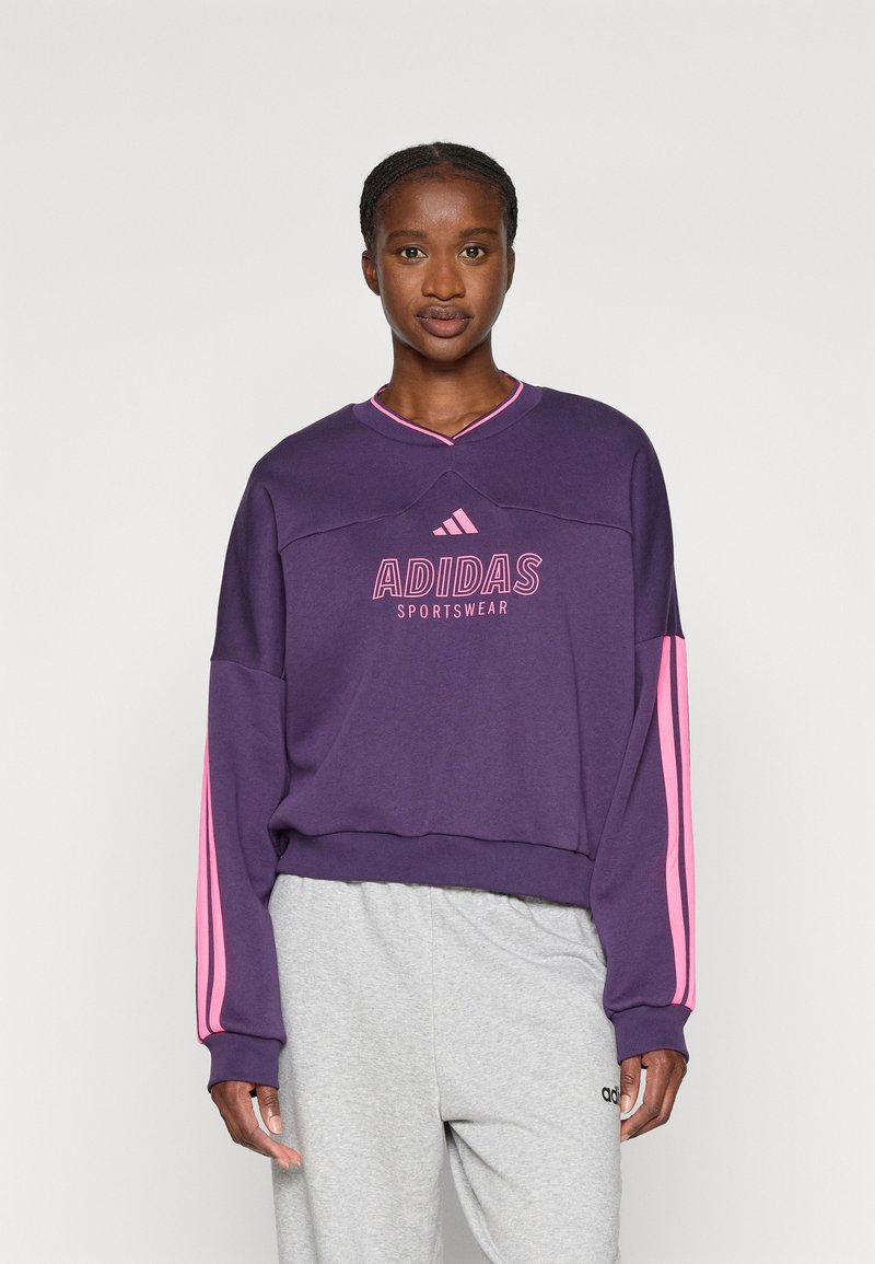 Vijolična dukserica s roza črtami ob rokavih, ki na sprednji strani nosi logotip "ADIDAS SPORTSWEAR" v roza barvi. Lahka tkanina.