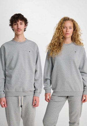 Jonge man en vrouw staan zij aan zij, gekleed in bijpassende grijze sweatshirts en joggingbroeken met kleine borstlogo's, neutrale gezichtsuitdrukkingen.