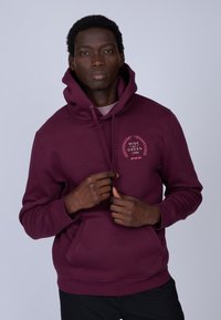 Haut à capuche bordeaux en tissu doux, avec une poche kangourou et un logo circulaire en rose avec des feuilles sur la poitrine. Capuche avec cordons incluse.