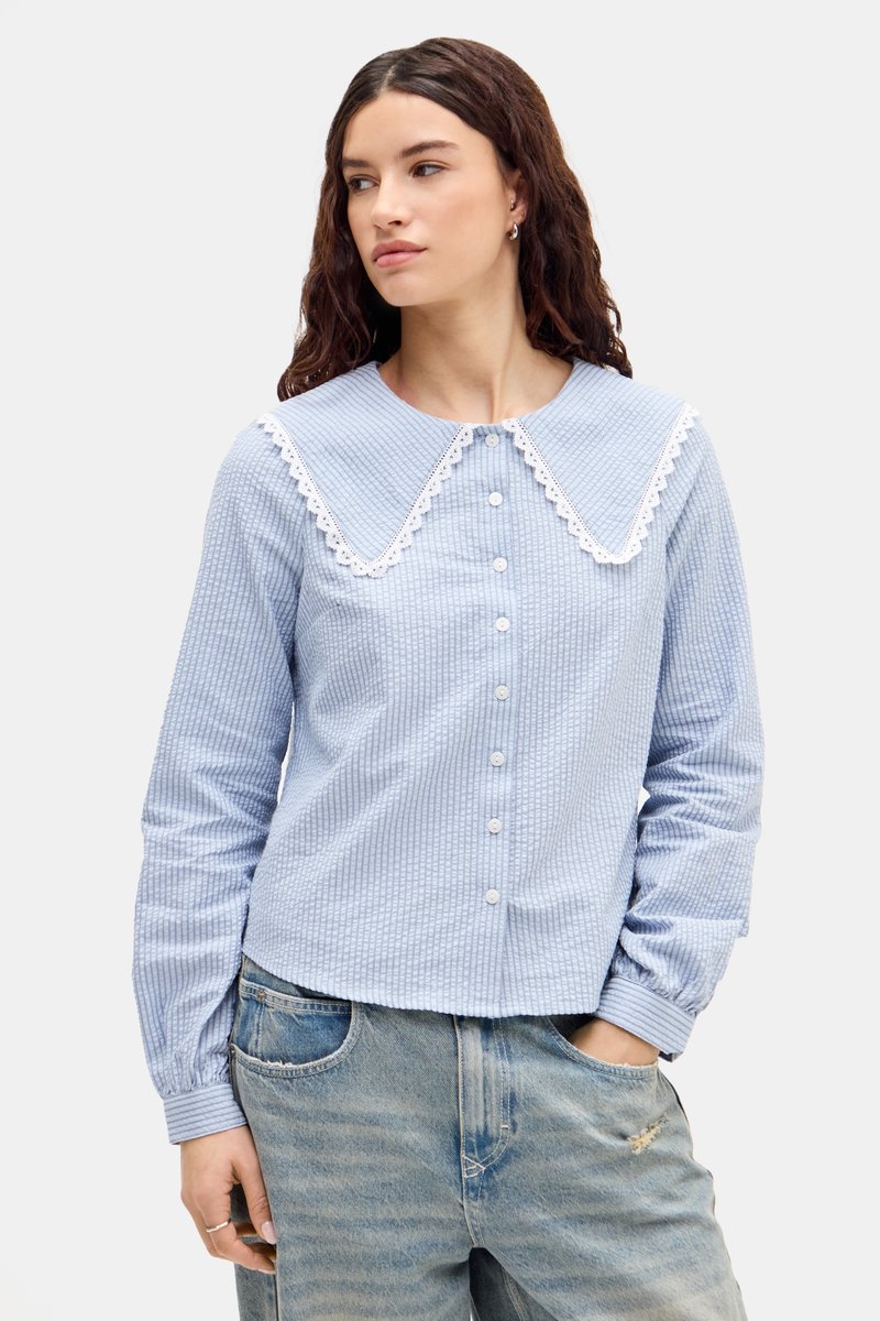 Camicia button-up azzurro chiaro, con texture e un ampio colletto in pizzo bianco, maniche lunghe e orlo dritto, abbinata a jeans blu sbiaditi.