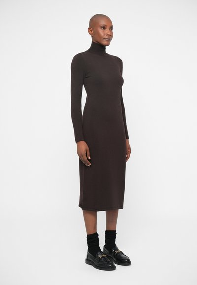 Rjava turtleneck obleka z dolgimi rokavi iz mehkega materiala, midi dolžine, telesu prilagojena zasnova. Nosi se z črnimi loafers in rebrastimi nogavicami.