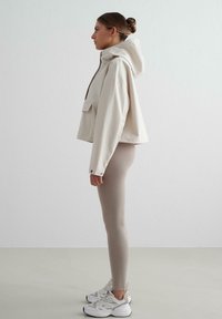 Haut à capuche blanc coupé avec une poche avant, associé à des leggings beiges et des baskets blanches, sur fond clair uni.