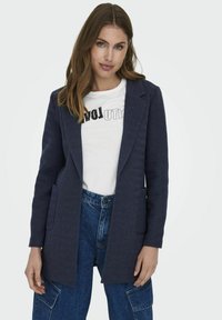 Sacou bleumarin cu model herringbone, cu două buzunare frontale, revere ascuțite și o croială ajustată, purtat peste un tricou grafic alb și pantaloni din denim blue.