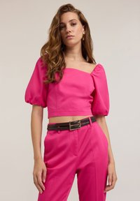 Top cropped fucsia con maniche a palloncino corte, abbinato a pantaloni a vita alta coordinati e una cintura marrone con fibbia dorata.