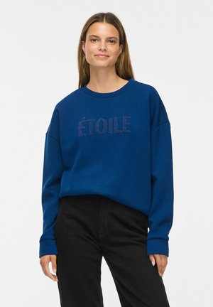 Kvinde med lige brunt hår iført en blå sweatshirt med teksten "ÉTOILE" og sorte bukser, stående mod en enkel hvid baggrund.